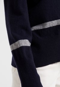 Marineblauer Pulloverärmel mit einem hellgrauen horizontalen Streifen über dem Handgelenk, getragen mit weißen Hosen.