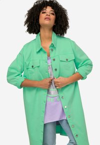 Giacca camicia oversized verde menta con chiusura a bottoni, due tasche sul petto e maniche arrotolate, indossata sopra una t-shirt lavanda.
