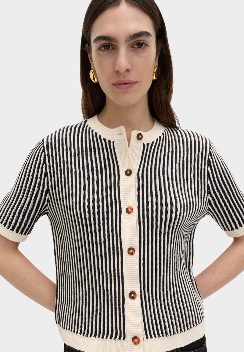 Cardigan à manches courtes rayé noir et blanc avec un col rond, doté de gros boutons marron et d'une texture côtelée le long des bords.