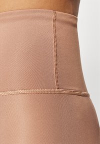 Vysoké, světlé hnědé legíny vyrobené z hladké, elastické tkaniny s vyztuženými švy a minimalistickým designem.