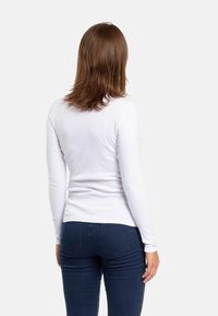 Camicia bianca a maniche lunghe con un design aderente, tessuto liscio e scollo rotondo, abbinata a jeans blu scuro con tasche posteriori.