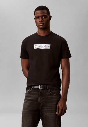 Mand iført sort Calvin Klein t-shirt og mørke jeans, stående foran en ensfarvet grå baggrund, vendt fremad med neutralt udtryk.