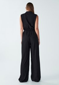 Mouwloze zwarte jumpsuit met een kraag, een cinched taille en wijde broekspijpen. De textuur is licht gekreukt en de stof lijkt lichtgewicht te zijn.