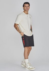 T-shirt crème à manches courtes avec logo, associée à un short cargo noir avec des zips rouges. Des baskets claires et des chaussettes blanches complètent le look.