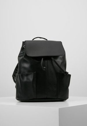 Black leather backpack with flap top, drawstring closure, front side pockets, and a handle, placed on a white pedestal against a light gray background.