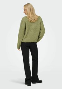 Groene, getextureerde trui met een losse pasvorm en een rafelige zoom, gecombineerd met zwarte broek en schoenen. Achteraanzicht weergegeven tegen een effen achtergrond.
