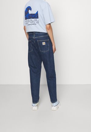 Osoba ubrana w niebieski t-shirt z nadrukiem i ciemnoniebieskie dżinsy z naszywką na tylnej kieszeni stoi na jednolitym jasnym tle.