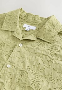 Hellgrünes Poloshirt mit gestickten Wirbelmustern, weißen Knöpfen und einem Pflegeetikett im Kragen.
