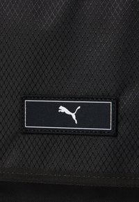 Tissu noir texturé avec un motif en losange et un logo Puma blanc sur une étiquette noire, cousue à la surface.