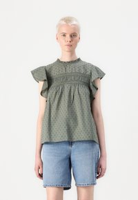 Grøn bomuldsbluse med flæseærmer, samlet bodice, prikmønster og høj halsudskæring. Sættes sammen med denimshorts.