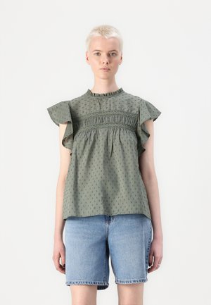 Blouse en coton vert avec des manches volantes, un buste froncé, une texture à pois et un col montant. Associée à un short en denim.