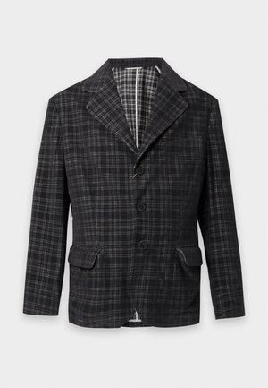 Jeune homme portant un blazer à carreaux foncé, un pull noir, un pantalon marron et des chaussures en cuir noires, debout devant un fond clair uni.