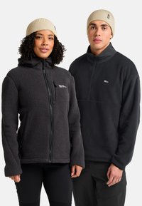 Due persone che indossano cappelli beige e giacche in pile nere, in piedi fianco a fianco su uno sfondo bianco.