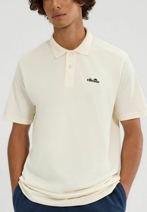 Młoda osoba ubrana w kremową koszulkę polo z krótkim rękawem z logo Ellesse na lewej piersi, z rękami w kieszeniach ciemnoniebieskich spodni.