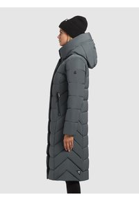 Femme portant un bonnet noir et un long manteau matelassé gris foncé avec une capuche, tournée vers la gauche sur un fond gris clair uni.