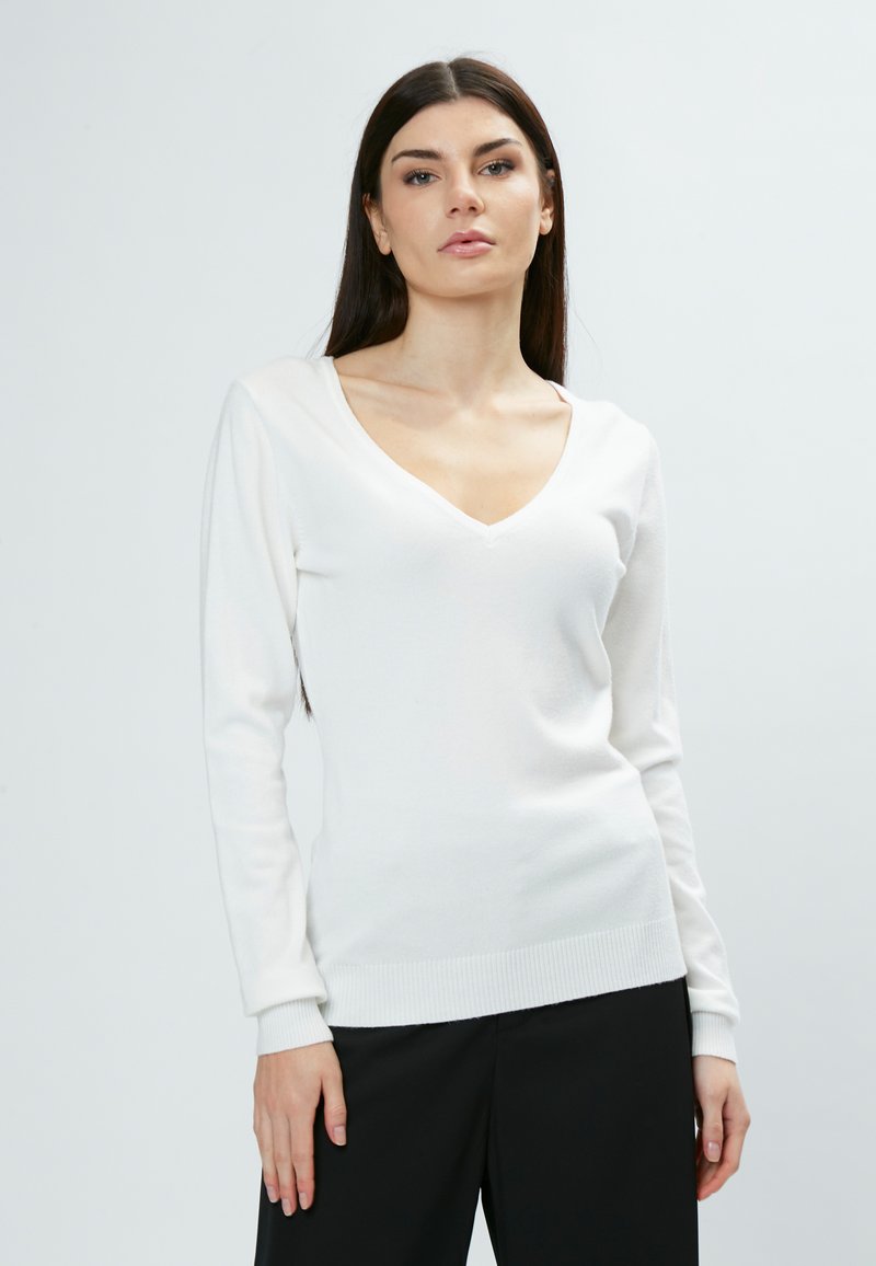 Weißer, langärmeliger V-Ausschnitt-Pullover aus weichem Material, mit taillierter Passform und gerippten Bündchen und Saum. Kombiniert mit schwarzen Hosen.