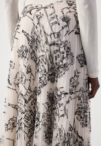 Plisseerok in crème met zwarte abstracte stadsafbeeldingen, gedragen met een lange mouw witte top, getoond van de taille naar beneden.