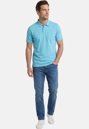 Mann steht mit einer Hand in der Tasche, trägt ein hellblaues Poloshirt, blaue Jeans und weiße Turnschuhe vor einfarbigem Hintergrund.