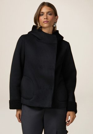 Manteau noir avec un col haut, tissu texturé, deux poches rondes et accents de bord en peluche. Le modèle le porte par-dessus un pantalon foncé.