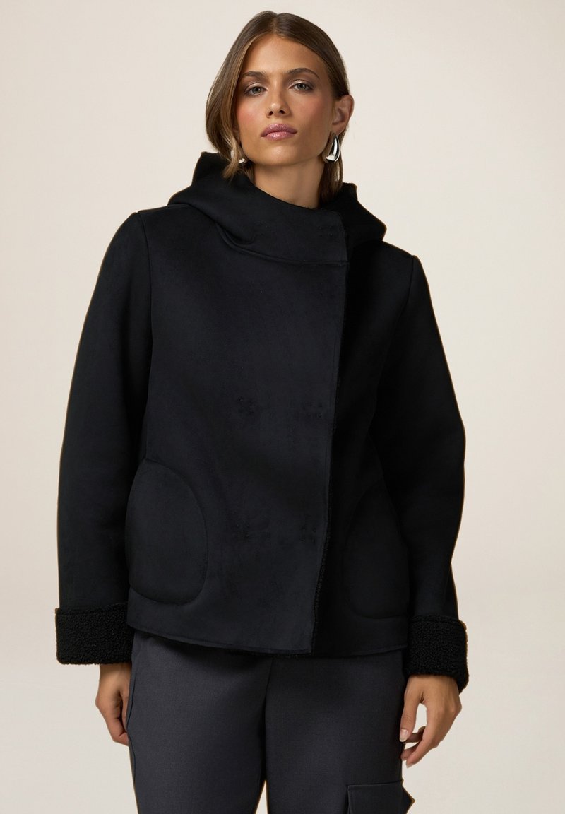 Cappotto nero con colletto alto, tessuto testurizzato, due tasche rotonde e dettagli in pelliccia sul fondo. Il modello lo indossa sopra pantaloni scuri.