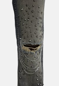 Donkergrijze denim broekspijp met gescheurd gat, versierd met metalen studs en hangende zilveren kettingen.