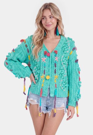 Kvinde med langt blondt hår bærer en turkis chunky strikket sweater med farverige pomponer og broderi, kombineret med revnede denimshorts.