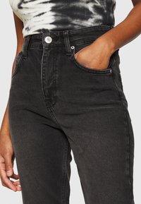 Jeans de mezclilla negros de talle alto con cierre de botón, bolsillos frontales y un acabado texturizado. Presenta un ajuste entallado y un diseño clásico.