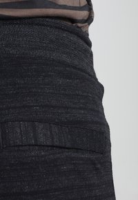 Gros plan sur une personne portant un haut rayé marron et noir rentré dans un pantalon taille haute gris foncé texturé avec un détail de poche.