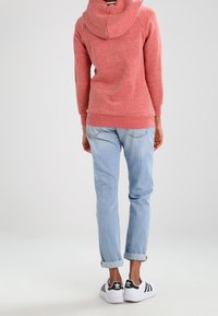 Sudadera con capucha de textura roja y capucha grande, combinada con vaqueros azul claro remangados y zapatillas blancas con rayas negras.