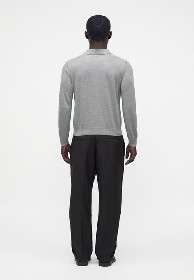Homme aux cheveux courts portant une chemise à manches longues gris clair et un pantalon noir, debout dos à un fond blanc uni.