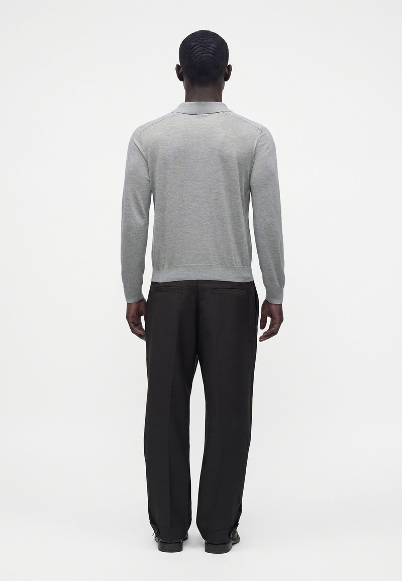 Homme aux cheveux courts portant une chemise à manches longues gris clair et un pantalon noir, debout dos à un fond blanc uni.