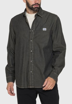 Uomo che indossa una camicia grigio scuro a maniche lunghe con bottoni sopra una maglietta bianca, in piedi davanti a uno sfondo chiaro e semplice.