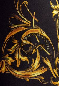 Černá látka s propracovanými zlatými barokními vzory a textem "VERSACE JEANS COUTURE" integrovaným do designu.