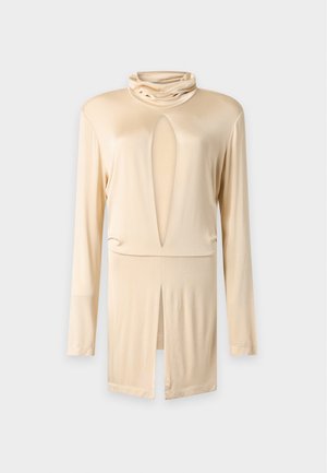 Femme aux longs cheveux foncés porte un haut beige à manches longues avec des découpes à l'avant, un pantalon large et des chaussures plates assorties, debout devant un fond uni.