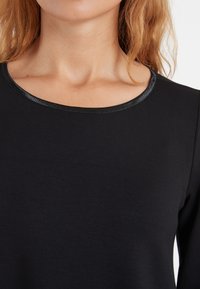 Schwarzes Oberteil mit rundem Ausschnitt, versehen mit einem glänzenden schwarzen Rand um den Kragen. Der Stoff wirkt glatt und leicht.