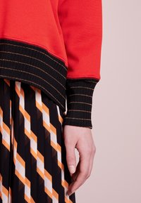 Suéter rojo con puños acanalados negros que presentan rayas doradas, combinado con una falda plisada de un patrón geométrico en blanco, negro y naranja.
