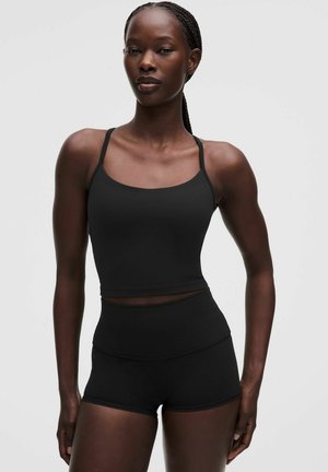 Mujer atlética con el pelo trenzado, vestida con una camiseta sin mangas negra ajustada y pantalones cortos de talle alto, de pie contra un fondo claro y liso.