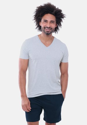 Mand med krøllet hår smiler, iført en lysegrå V-hals t-shirt og mørke shorts, stående med den ene hånd i lommen mod en ensfarvet baggrund.