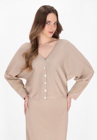 Béžový oversized cardigan s V-výstřihem, dlouhými rukávy a kovovými knoflíky vpředu; má texturovanou, jemnou látku.