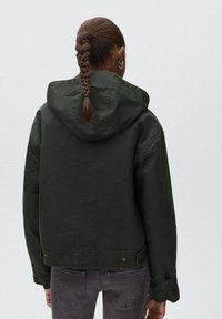 Mörkgrön huva jacka med avslappnad passform, synliga sömmar och tryckknappsdetaljer, kombinerad med grå jeans och en flätad frisyr.