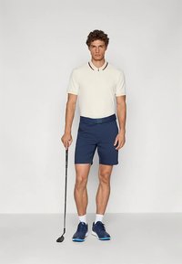 Biała koszulka polo z kołnierzykiem i bordowymi akcentami, zestawiona z granatowymi szortami. Model trzyma kij golfowy, ma na sobie ciemne buty sportowe i białe skarpetki.