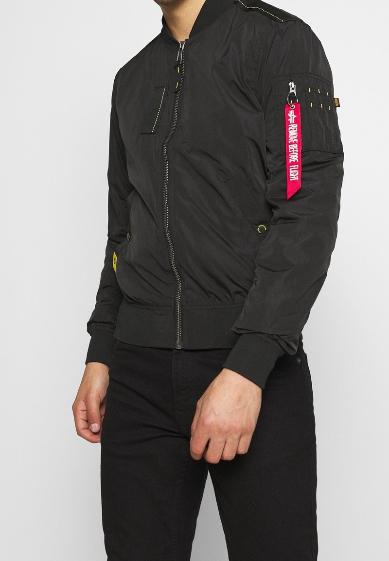 Homme portant une veste bomber noire avec une fermeture éclair à l'avant, des poignets côtelés et une étiquette rouge "Remove Before Flight" sur la poche de la manche gauche.