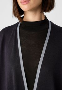 Ciemnoszary kardigan z szarym wykończeniem, nałożony na czarny golf. Miękka tekstura, otwarty fason i brak widocznych zapięć.