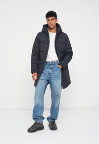 Mørkeblå puffjakke med hætte, der har et quiltet design. Sammen med lyseblå jeans og sorte sneakers. Enkel, afslappet stil.