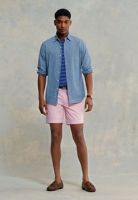 Svetlo modra chambray srajca preko mornarsko modre in bele črtaste polo majice, v kombinaciji s rožnatimi kratkimi hlačami in rjavimi čolnarskimi čevlji. Vključuje tudi zapestno uro.