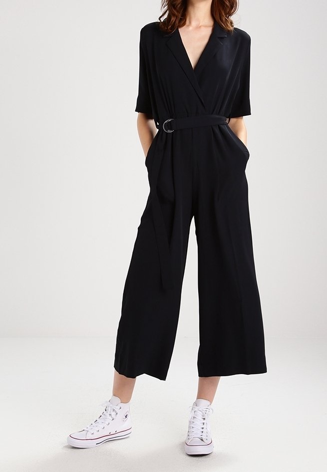 Schwarzer Jumpsuit mit kurzen Ärmeln, weitem Bein, tailliert mit einem Gürtel und seitlichen Taschen, kombiniert mit weißen High-Top-Sneakern. Glatte Textur.