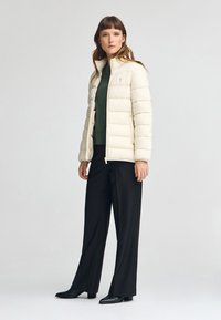 Puffjacka i off-white tyg, med hög krage och dragkedja. Modellen har på sig en grön topp och svarta byxor med platta svarta skor.