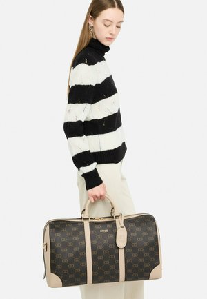 Jeune femme portant un pull à rayures noir et blanc, tenant un grand sac de voyage Liu Jo marron et beige à motifs par ses poignées.