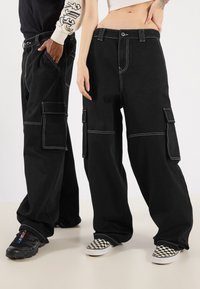 Black cargo pants featuring white stitching, large side pockets, and a loose fit, paired with diverse footwear styles.