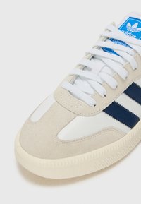 Beige mocka och vit läder sneaker med blå ränder, vita snörningar och strukturerad gummiyttersula. Har klassiska designelement.